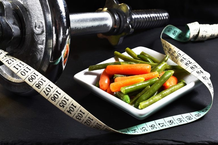 Still life of a metal dumbbell, a tape measure, and a dish of steamed carrots and green beans on a dark surface